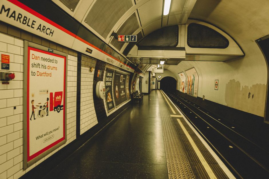 An empty platform inside Archway underground station, with white tiled walls and a curved ceiling. A large advertisement board on the left displays a red and white poster about shifting drums to Dartford, featuring illustrations of people and drums. The platform's edge has tactile paving strips for visually impaired passengers, and a dark, empty tunnel entrance is visible in the distance. Along the wall, there are black benches and various signs, including departure information and safety notices. The lighting is bright, evenly illuminating the scene, while the overall environment appears clean and organized. This setting, captured by Man With a Van Archway, illustrates the typical environment of a London tube station, where furniture or boxes can be loaded onto transport vehicles as part of home relocation or moving logistics.